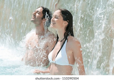 Happy smiling couple refresh under fresh waterfall at summer swimming pool