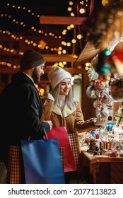 Happy Smiling Couple Shopping At Christmas Street Market, Choosing Gifts. Winter Holidays, Vacation, Travel, Purchase Conception. Outdoor Night Portrait
