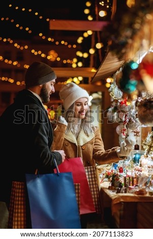 Happy smiling couple shopping at Christmas street market, choosing gifts. Winter holidays, vacation, travel, purchase conception. Outdoor night portrait
