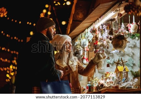 Happy smiling couple shopping at Christmas street market, choosing gifts. Winter holidays, vacation, travel, purchase conception. Outdoor night portrait. Copy, empty space for text
