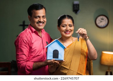 Happy smiling couple showing new house keys by toy home while looking camera - concept of new home purchasing, home loan and investment.