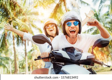 Happy smiling couple travelers riding motorbike scooter in safety helmets during tropical vacation under palm trees - Shutterstock ID 1576813492