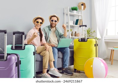 Happy smiling couple travelers sitting on sofa with suitcases at home booking tickets online via laptop. Young man and woman wearing beach hat getting ready for holiday trip. Vacation concept. - Powered by Shutterstock