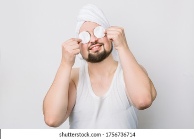 Happy Smiling Crazy Funny Bearded Man With White Towel Oh His Head, Holding Two Cotton Pads And Hiding His Eyes While Caring About His Skin. Close Up Portrait On White Background. Spa For Men