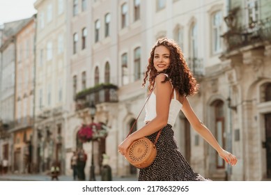 Happy smiling curly brunette woman wearing trendy summer outfit with round wicker shoulder rotan bag, white top, polka dot midi skirt, walking in street of European city. Fashion, lifestyle concept 