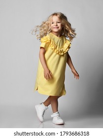 Happy Smiling Cute Blonde Curly Kid Girl Preschooler In Yellow Dress And Sneakers Walking Moving Over Grey Wall Background. Stylish Casual Fashion For Children