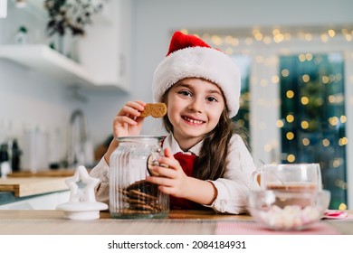 Happy Smiling Cute Little Girl Kid In Santa Claus Hat Sitting On The Decorated For Winter Holidays Modern Kitchen And Eating Gingerbread Cookies From The Jar With Cocoa Drink. Festive Season