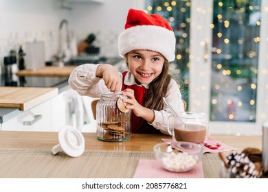 Happy Smiling Cute Little Girl Kid In Santa Claus Hat Sitting On The Decorated For Winter Holidays Modern Kitchen And Reaching Gingerbread Cookies From The Jar With A Cocoa Drink. Festive Season