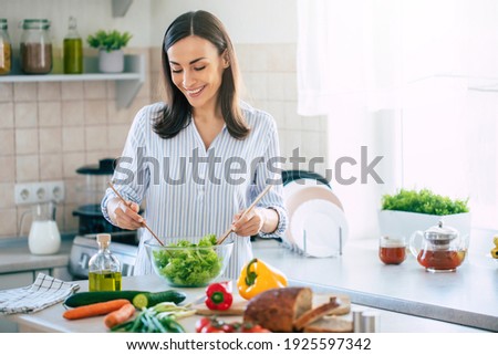 Happy smiling cute woman is preparing a fresh healthy vegan salad with many vegetables in the kitchen at home and trying a new recipe