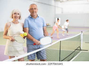 Happy smiling elderly couple, man and woman in sportswear with rackets and balls in hands posing near net on indoor pickleball court after friendly match - Powered by Shutterstock