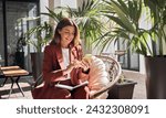 Happy smiling elegant mature middle aged business woman wearing suit holding cellphone using mobile cell phone looking at smartphone sitting in comfortable chair in sunny office with green plants.