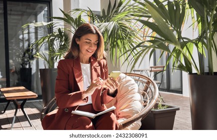 Happy smiling elegant mature middle aged business woman wearing suit holding cellphone using mobile cell phone looking at smartphone sitting in comfortable chair in sunny office with green plants. - Powered by Shutterstock