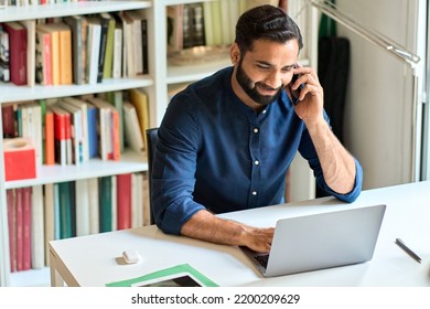 Happy smiling ethnic eastern indian professional business man sitting at work desk talking on cell phone, eastern businessman making mobile phone call by cellphone working at home or in office.