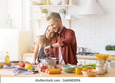 Happy Smiling Family Couple Cooking On Loft Kitchen. Young Attractive Wife Or Girlfriend Hugging Loving Husband Or Boyfriend While Man Preparing Dinner From Fresh Vegetable. Couple Making Organic Food