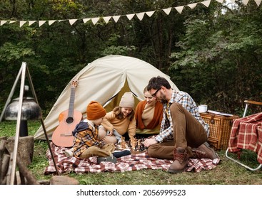 Happy smiling family playing chess game during camping trip in nature, parents with kids playing board games together while sitting on checkered plaid near tent at campsite, enjoying outdoor tourism - Powered by Shutterstock