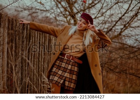 Happy smiling fashionable blonde woman wearing trendy autumn outfit with classic brown coat, trendy cap, beige turtleneck sweater, leather belt, plaid mini skirt, posing outdoor. Copy, empty space 