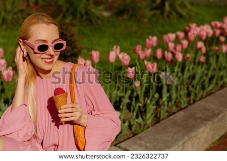 Happy smiling fashionable blonde woman wearing trendy pink sunglasses, pleated dress, holding fruit ice cream, posing in street of European city. Copy, empty space for text