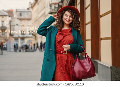 Happy smiling fashionable curvy woman wearing trendy autumn outfit: orange hat, snakeskin print dress, belt, green coat, holding red wicker leather bag, posing in street of European city. Copy space