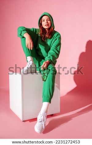 Happy smiling fashionable freckled redhead girl wearing trendy green suit with hoodie, joggers,  white sneakers, sitting, posing on pink background. Full-length studio portrait
