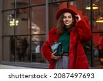 Happy smiling fashionable woman wearing trendy orange faux fur coat, hat, green turtleneck sweater, holding stylish bag, posing in street of city. Outdoor winter fashion portrait. Copy, empty space 