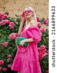 Happy smiling fashionable woman wearing trendy summer fuchsia color dress, pink sunglasses, holding stylish green faux leather bag, posing near blooming roses