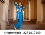 Happy smiling fashionable woman wearing trendy outfit with blue  coat, turtleneck, wide leg trousers, sunglasses, white boots, bag, posing in street of European city. Full-length outdoor portrait