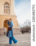 Happy smiling fashionable woman wearing trendy blue coat, wide leg trousers, white boots, burgundy leather shoulder bag, walking in street of European city. Full-length outdoor portrait