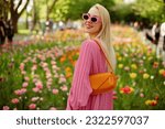 Happy smiling fashionable woman wearing trendy pink sunglasses, pleated dress, with orange faux patent leather baguette bag, posing among flowers. Copy, empty space for text
