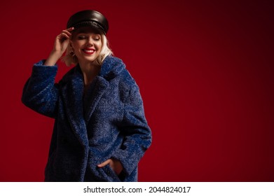 Happy smiling fashionable woman wearing trendy blue faux fur coat, leather cap, posing in studio, on red background. Copy, empty space for text 