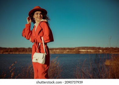 Happy Smiling Fashionable Woman Wearing Trendy Orange Trench Coat, Hat, Holding White Textured Leather Bag, Posing In Nature. Outdoor Autumn Fashion Portrait. Copy, Empty Space For Text