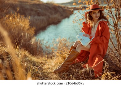 Happy smiling fashionable woman wearing trendy orange color hat, autumn trench coat, brown high boots, with white faux leather bag, posing in nature. Copy, empty space for text