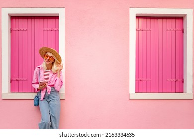 Happy smiling fashionable woman wearing stylish summer outfit holding, using her smartphone, posing near pink color house. Copy, empty space for text

 - Powered by Shutterstock