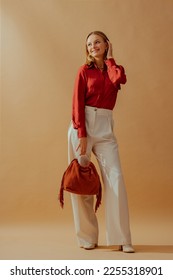 Happy smiling fashionable woman wearing elegant satin blouse, white wide leg trousers, holding trendy suede fringed bag, posing on beige background. Full-length studio fashion portrait - Shutterstock ID 2255318901