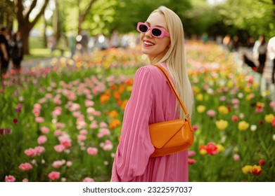 Happy smiling fashionable woman wearing trendy pink sunglasses, pleated dress, with orange faux patent leather baguette bag, posing among flowers. Copy, empty space for text - Shutterstock ID 2322597037