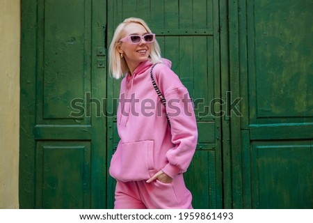 Happy smiling fashionable woman wearing trendy pink sport chic style outfit, sunglasses, posing in street on green background. Glasses, pink hoodie, trousers. Spring fashion concept. Copy, empty space