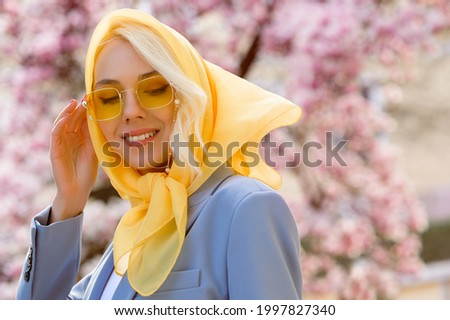 Happy smiling fashionable woman wearing trendy yellow headscarf, sunglasses, posing near blooming magnolia tree. Spring fashion, lifestyle conception. Outdoor portrait. Copy, empty space for text
