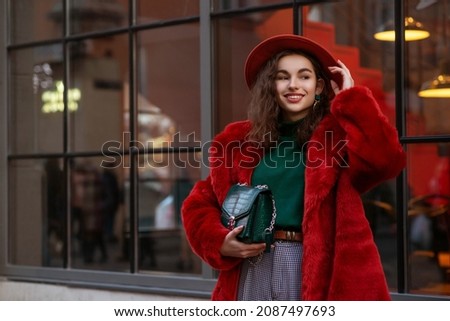 Happy smiling fashionable woman wearing trendy orange faux fur coat, hat, green turtleneck sweater, holding stylish bag, posing in street of city. Outdoor winter fashion portrait. Copy, empty space 