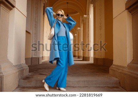 Happy smiling fashionable woman wearing trendy outfit with blue  coat, turtleneck, wide leg trousers, sunglasses, white boots, bag, posing in street of European city. Full-length outdoor portrait
