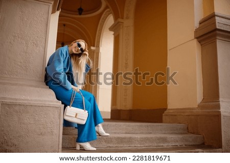 Happy smiling fashionable woman wearing trendy outfit with blue  coat, wide leg trousers, sunglasses, white ankle boots, quilted leather bag, posing in street. Full-length outdoor portrait. Copy space