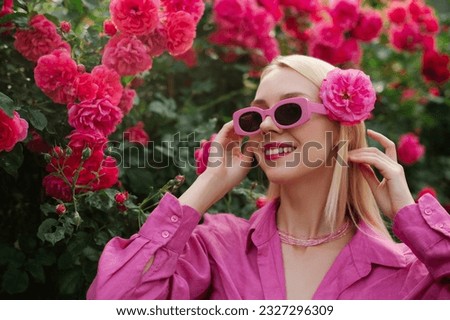 Happy smiling fashionable woman wearing trendy pink sunglasses, linen summer shirt, posing in garden, among flowers. Copy, empty space for text
