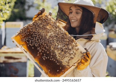 Happy smiling female Beekeeper in protective suit holding honeybee frame with bees at apiary - Powered by Shutterstock