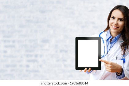 Happy smiling female doctor pointing at tablet pc with empty copy space area for slogan or text over office white brick loft wall. Medical call center concept. Zoom online video consult. iPad touchpad