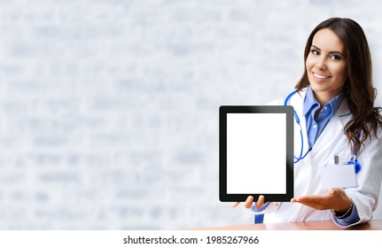Happy smiling female doctor showing tablet pc iPad touchpad with blank copy space area for slogan or text, over office white brick loft wall. Medical call center concept. Zoom, Skype video conference.