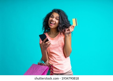 Happy smiling female fashion blogger with a bunch of shopping bags holding credit card and making online purchases using her mobile phone. Studio shot isolated over blue background.