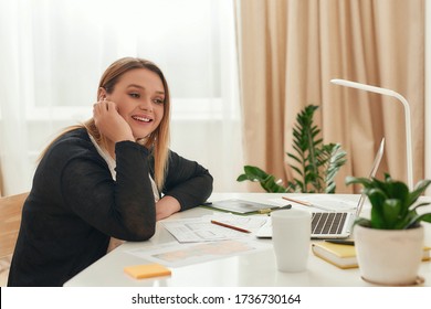 Happy Smiling Female Interior Designer Or Architect Wearing Wireless Headphones And Talking To Someone While Sitting At Her Workplace, Working From Home. Remote Work. Positive Human Emotions