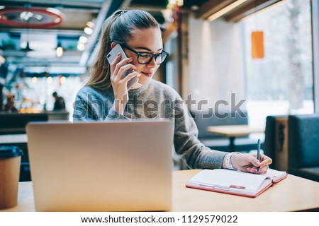 Happy smiling female journalist planning meeting while talking about interview via application on mobile phone and enjoying distance job, successful programmer calling to customer service indoors