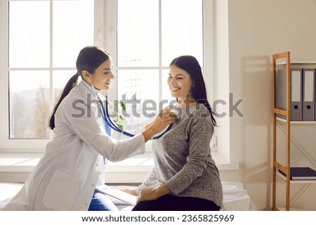 Happy smiling female patient in clinic for a medical checkup. Yong friendly physician doctor with stethoscope examining woman's lungs, breathing and heartbeat at the hospital in exam room.