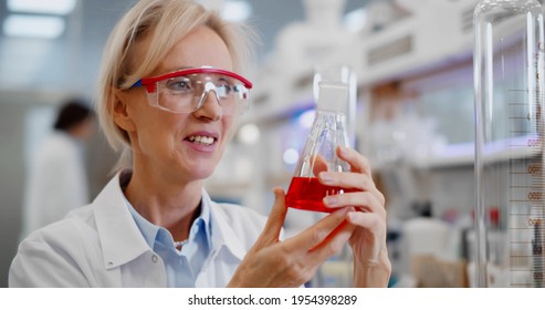Happy smiling female scientist in goggles with chemicals in flask making test or research in laboratory. Mature woman chemist holding glassware with red liquid smiling 