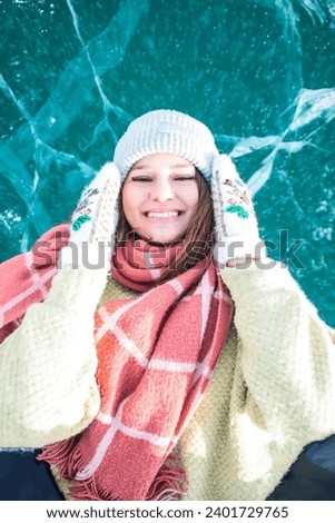 Happy smiling female traveler dressed in a warm winter hat and mittens lying on frozen Baikal lake with cracked ice floes 