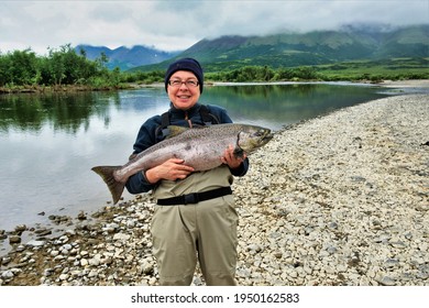 A happy, smiling fisherwoman holds a large King salmon that she caught after a long fight.  She is standing on a rocky bank of a remote river in the mountains of Alaska.